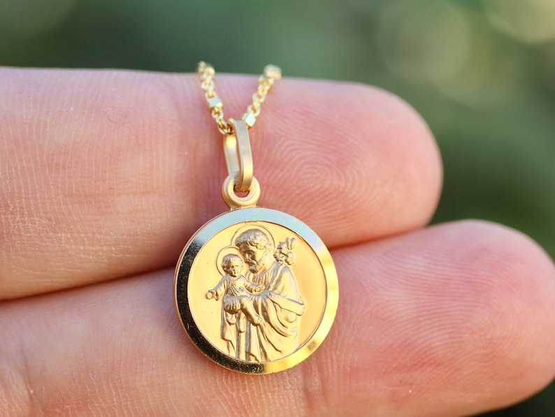 Pode incluir: Colar com pingente de ouro que apresenta um amuleto redondo com uma imagem detalhada de S&atilde;o Jos&eacute; segurando o menino Jesus. O amuleto tem uma borda elevada e um acabamento liso e polido.