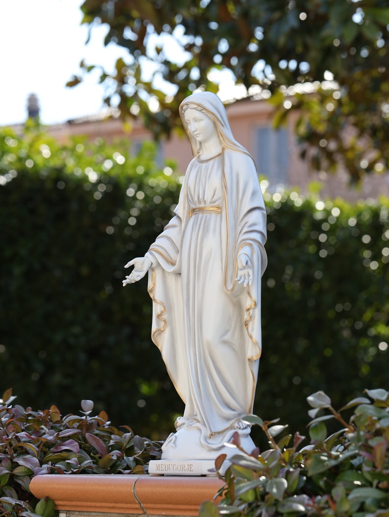 May include: White statue of the Virgin Mary with a gold trim on her robe. The statue is standing on a base with the word "Medjugorje" inscribed on it.