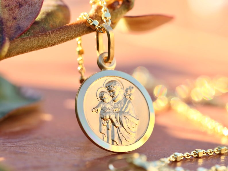 Pode incluir: Colar com pingente de ouro que apresenta uma imagem detalhada de Santo Ant&ocirc;nio de P&aacute;dua segurando um beb&ecirc; Jesus. O pingente &eacute; redondo e tem um acabamento liso e polido.
