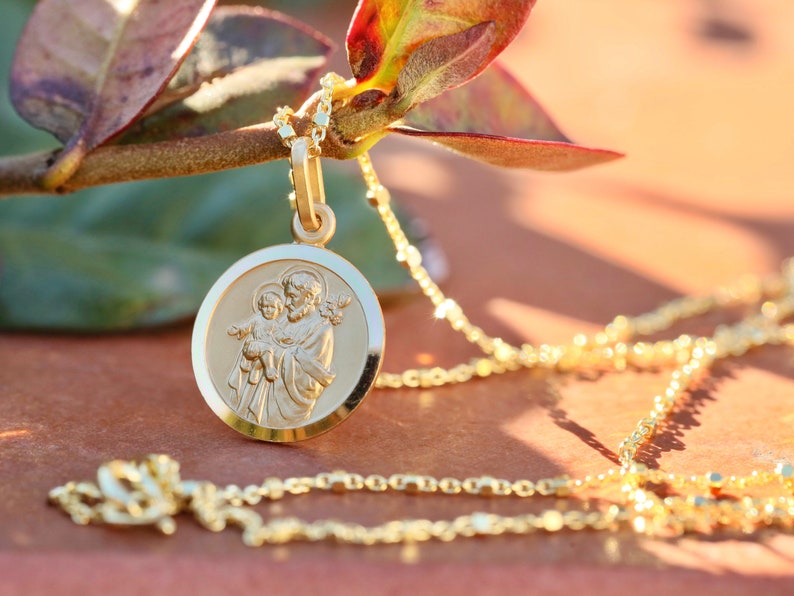 Pode incluir: Um colar de corrente de ouro com um pingente redondo que apresenta uma imagem detalhada de S&atilde;o Jos&eacute; segurando o menino Jesus. O pingente tem uma borda elevada e um acabamento liso e polido.