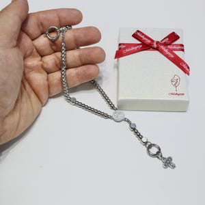 May include: A silver rosary necklace with a round pendant featuring a design of small white stones. The rosary is on a hand and a white box with a red ribbon and the text "Mahogany" is in the background.