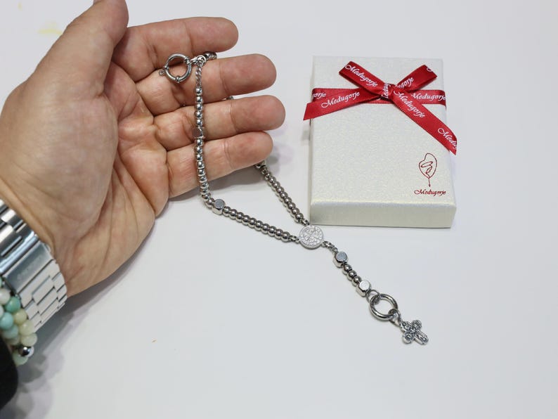May include: A silver rosary necklace with a round pendant featuring a design of small white stones. The rosary is on a hand and a white box with a red ribbon and the text "Mahogany" is in the background.