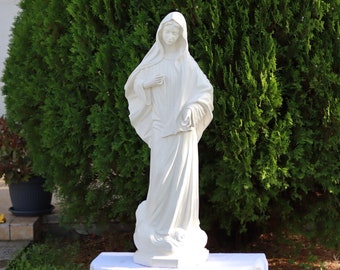 May include: White statue of the Virgin Mary standing on a pedestal. The statue is outdoors in front of a green bush.