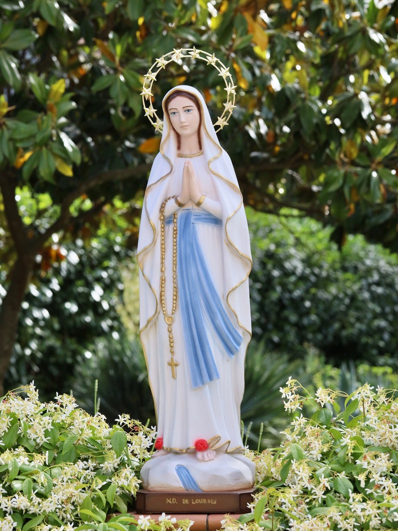 May include: A white statue of the Virgin Mary, wearing a blue sash and a crown of stars, stands on a pedestal with the words "N.D. de Lourdes". The statue is surrounded by white flowers.