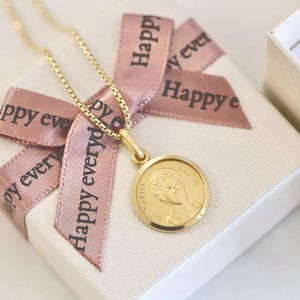 Peut inclure: Un collier pendentif en or avec un charm rond représentant un portrait de profil. Le collier repose sur un coffret cadeau blanc noué avec un ruban rose imprimé avec "Happy everyday" en noir. La chaîne est délicate.