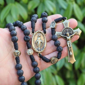Peut inclure: Un chapelet noir avec des perles en pierre de lave, un crucifix doré et un pendentif ovale doré représentant une figure religieuse. Le chapelet est présenté sur un fond de feuillage vert.