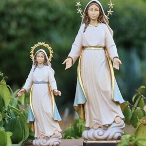 K&ouml;nnte beinhalten: Zwei wei&szlig;e Holzstatuen der Jungfrau Maria, eine gr&ouml;&szlig;er als die andere. Beide Statuen tragen wei&szlig;e Gew&auml;nder mit blauen B&auml;ndern und goldenen Kronen. Die gr&ouml;&szlig;ere Statue hat einen goldenen Sternenhalo, w&auml;hrend die kleinere Statue eine goldene Krone mit Sternen tr&auml;gt. Beide Statuen stehen auf Wolken.