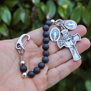Könnte beinhalten: Schwarze Perlenkette mit einem silbernen Kruzifix, einem silbernen Anhänger der Jungfrau Maria und einem silbernen Anhänger mit der Aufschrift "INRI".