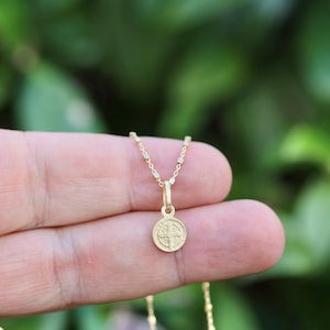 Può includere: Una collana dorata con un piccolo pendente rotondo con un design dettagliato. Il pendente è appeso a una catena sottile. La collana è tenuta contro uno sfondo verde sfocato, suggerendo un ambiente esterno. Il pendente ha un tema religioso.