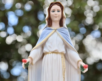Könnte beinhalten: Eine weiße Statue der Jungfrau Maria mit einem blauen Gewand und goldenen Verzierungen. Sie hält in jeder Hand einen Strauß roter Rosen. Die Statue steht vor einem verschwommenen Hintergrund aus grünem Laub.