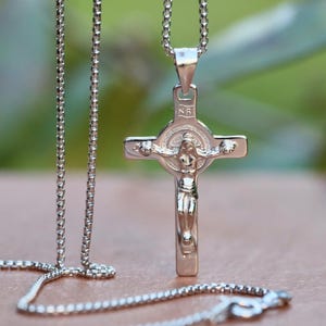 Può includere: Una collana a catena d'argento con un ciondolo d'argento che presenta un crocifisso con una figura di Gesù Cristo. Il ciondolo ha un design circolare con un bordo rialzato attorno alla figura.