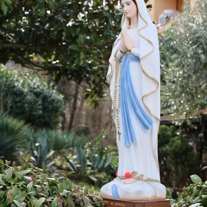 May include: A white statue of the Virgin Mary, wearing a blue sash and a white veil, stands on a pedestal with the words "Notre Dame de Lourdes" inscribed on it. The statue is surrounded by green foliage.