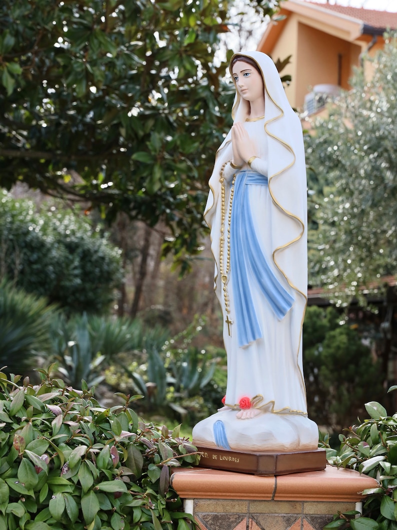May include: A white statue of the Virgin Mary, wearing a blue sash and a white veil, stands on a pedestal with the words "Notre Dame de Lourdes" inscribed on it. The statue is surrounded by green foliage.