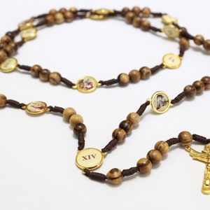 Puede incluir: Un rosario de madera marrón con detalles dorados y una cruz dorada. Las cuentas están enhebradas en un cordón marrón. El rosario tiene pequeños medallones con imágenes. Los medallones tienen el número XIV.