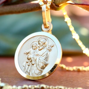 Pode incluir: Um colar com um pingente redondo dourado que apresenta uma imagem em relevo de S&atilde;o Jos&eacute; segurando o menino Jesus. O pingente est&aacute; preso a uma delicada corrente de ouro.