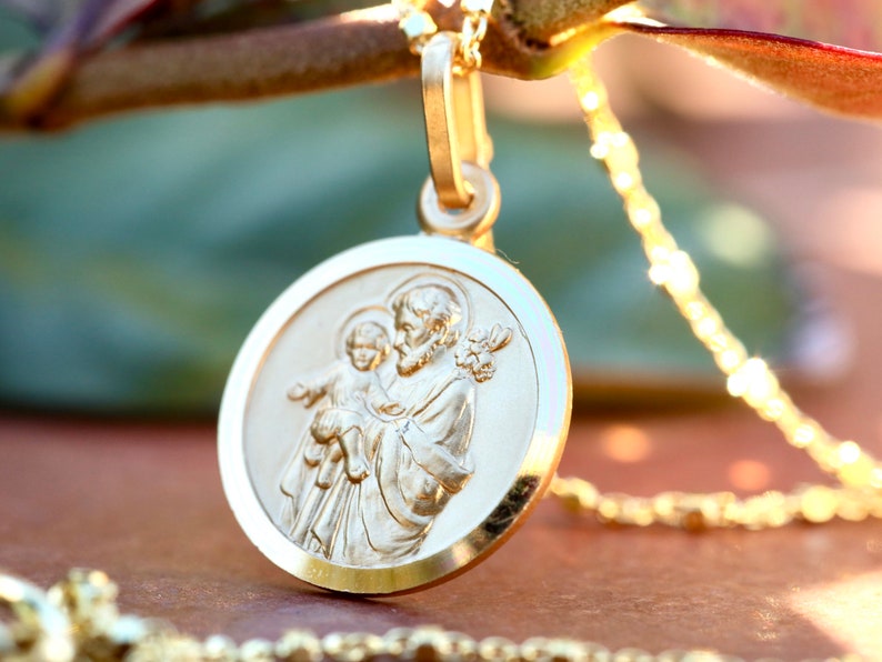 Pode incluir: Um colar com um pingente redondo dourado que apresenta uma imagem em relevo de S&atilde;o Jos&eacute; segurando o menino Jesus. O pingente est&aacute; preso a uma delicada corrente de ouro.