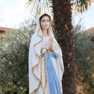 May include: A white statue of the Virgin Mary with a blue robe and a gold rosary. The statue is standing in front of a palm tree and a building.