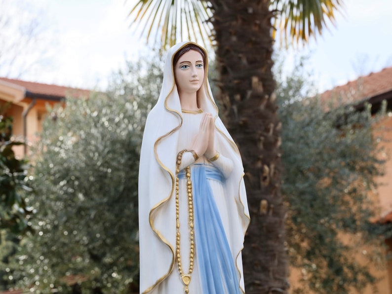 May include: A white statue of the Virgin Mary with a blue robe and a gold rosary. The statue is standing in front of a palm tree and a building.