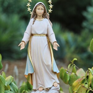 K&ouml;nnte beinhalten: Eine wei&szlig;e Statue der Jungfrau Maria mit einem goldenen Sternenhalo. Sie steht auf einer Wolke und hat ihre Arme ausgestreckt. Die Statue steht auf einem braunen Sockel.