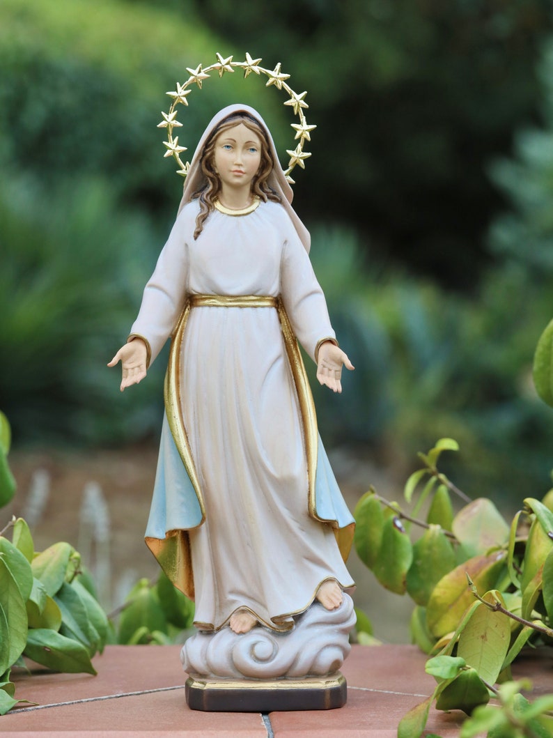 K&ouml;nnte beinhalten: Eine wei&szlig;e Statue der Jungfrau Maria mit einem goldenen Sternenhalo. Sie steht auf einer Wolke und hat ihre Arme ausgestreckt. Die Statue steht auf einem braunen Sockel.
