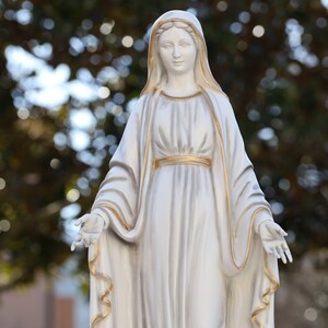 May include: White statue of the Virgin Mary with a gold trim on her robe and a soft, blurred background.