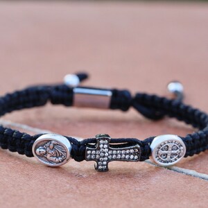 May include: A black braided bracelet with a silver cross charm, a silver oval charm with a religious figure, and a silver oval charm with a religious symbol.