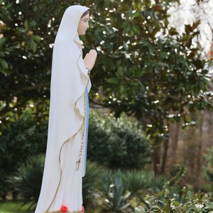 May include: White statue of the Virgin Mary with a blue robe and a rosary. The statue is standing on a white base with red flowers. The statue is in front of a green bush.