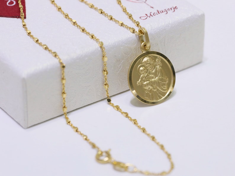Pode incluir: Um colar de corrente de ouro com um pingente redondo que apresenta uma imagem detalhada de S&atilde;o Jos&eacute; segurando o menino Jesus. O pingente est&aacute; sobre uma caixa branca com a palavra "Medjugorje" escrita nela.