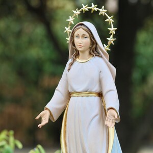 K&ouml;nnte beinhalten: Eine wei&szlig;e Statue einer Frau mit einem goldenen Sternenhalo, die ein wei&szlig;es Gewand mit einem goldenen G&uuml;rtel tr&auml;gt. Die Statue steht mit ausgestreckten Armen.