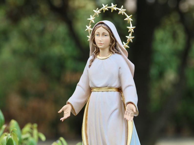 K&ouml;nnte beinhalten: Eine wei&szlig;e Statue einer Frau mit einem goldenen Sternenhalo, die ein wei&szlig;es Gewand mit einem goldenen G&uuml;rtel tr&auml;gt. Die Statue steht mit ausgestreckten Armen.
