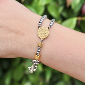 Pode incluir: Uma pulseira de rosário dourada e prateada. A pulseira apresenta um medalhão dourado com um design em relevo, contas douradas e prateadas e um pequeno pendente de cruz. A pulseira é usada num pulso.