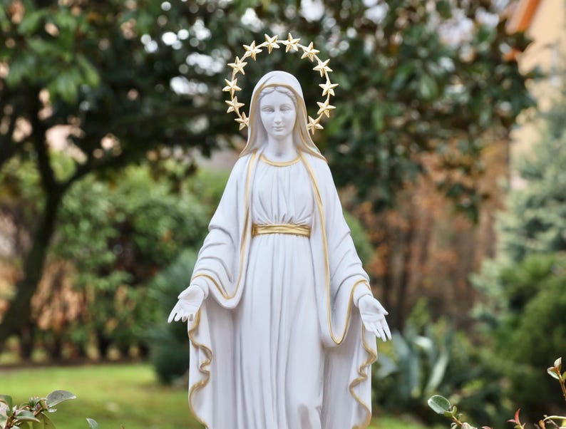 May include: White statue of the Virgin Mary with a gold star halo. The statue is standing in a garden setting.