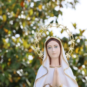 May include: A white statue of the Virgin Mary with a gold star halo. She is wearing a white robe and has her hands clasped in prayer.