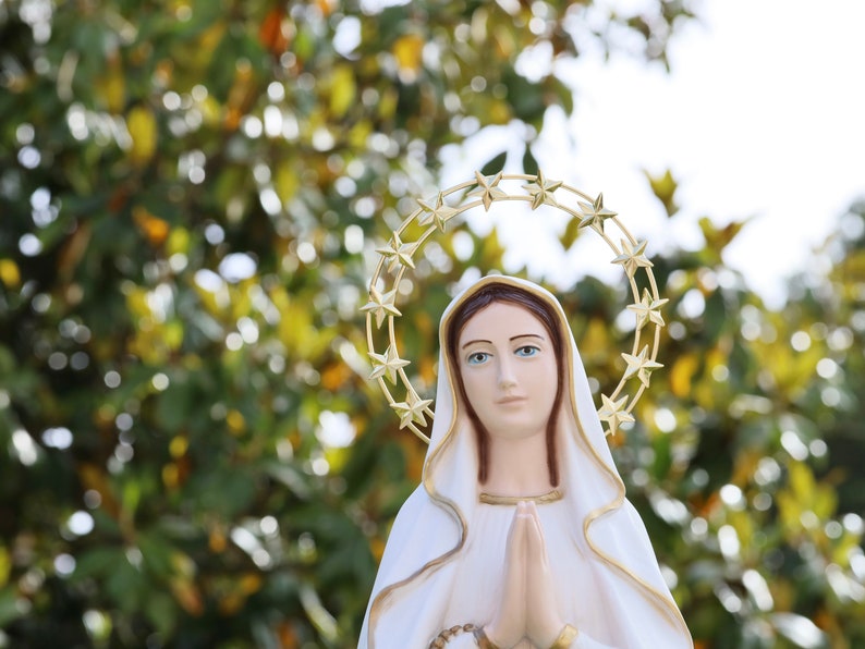 May include: A white statue of the Virgin Mary with a gold star halo. She is wearing a white robe and has her hands clasped in prayer.