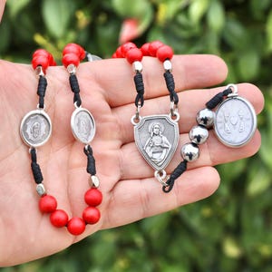 May include: A rosary featuring red beads, silver-coloured metal components, and black cord. The rosary includes religious medallions with detailed imagery. The background is a blurred green.