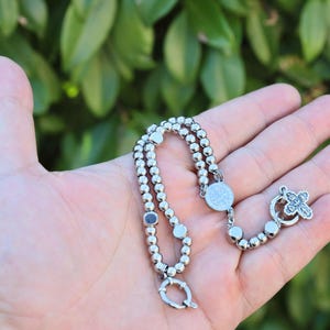 May include: A silver beaded bracelet with a silver cross charm and a round silver pendant. The bracelet is on a hand.
