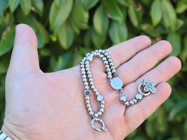 May include: A silver beaded bracelet with a silver cross charm and a round silver pendant. The bracelet is on a hand.