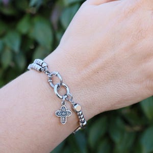 May include: A silver bracelet with a silver cross charm and a silver chain. The bracelet is on a person's wrist.