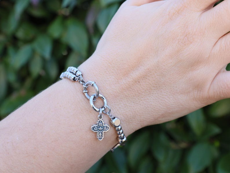 May include: A silver bracelet with a silver cross charm and a silver chain. The bracelet is on a person's wrist.