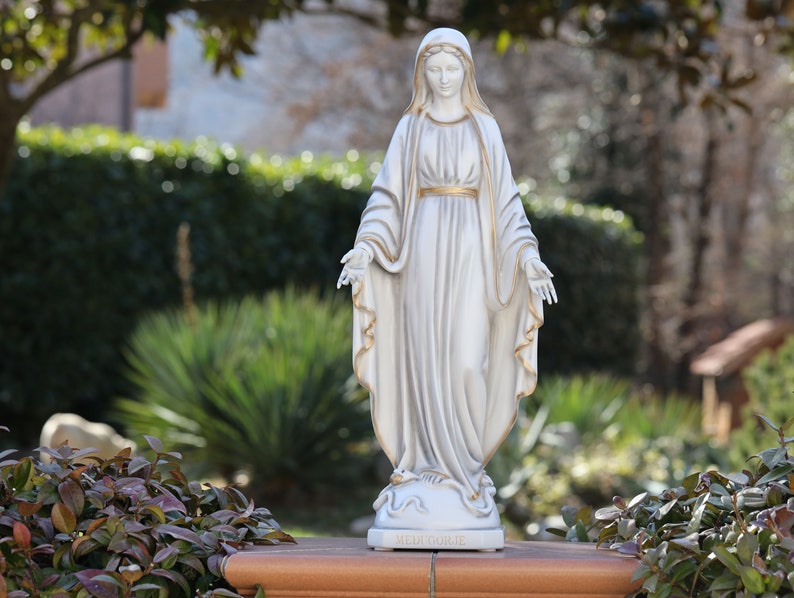 May include: White statue of the Virgin Mary with a gold trim on her robe. The statue is standing on a base with the words "MEDJUGORJE" inscribed on it.