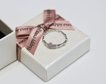 May include: A silver ring with a small cluster of stones is displayed on a white gift box tied with a pink ribbon. The ribbon has the words "Happy everyday" printed on it. The ring is the focal point, resting on the box.