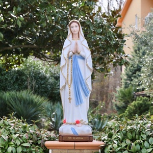 May include: A white statue of the Virgin Mary, wearing a blue robe and holding a rosary. The statue is standing on a pedestal with the words "Ave Maria" inscribed on it. The statue is surrounded by green foliage.