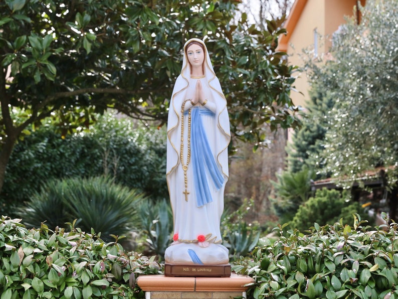 May include: A white statue of the Virgin Mary, wearing a blue robe and holding a rosary. The statue is standing on a pedestal with the words "Ave Maria" inscribed on it. The statue is surrounded by green foliage.