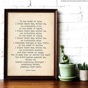 Op de afbeelding: Een ingelijste print met een donkerbruine rand toont een citaat van Albert Camus op een crèmekleurige achtergrond. De tekst is in een klassiek schreeflettertype. Een kleine groene potplant en een cactus zijn ook zichtbaar.