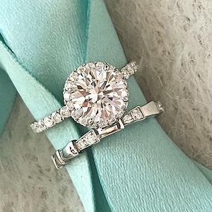 May include: A close-up of a silver engagement ring with a large, round, clear diamond center stone. The ring is set with smaller diamonds around the center stone and on the band. The ring is on a light blue background.