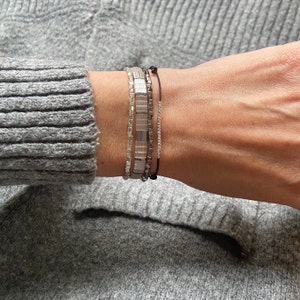 Op de afbeelding: Een stapel zilveren en witte kralen armbanden op de pols van een persoon. De armbanden zijn gelaagd en bevatten een dun zwart touwtje armband met een kleine zwarte kraal.