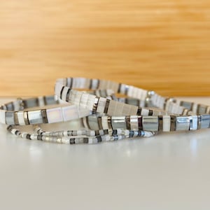 May include: Four bracelets stacked on a white surface. The bracelets are made of white and grey beads. The top bracelet is white with a silver stripe. The second bracelet is grey with a white stripe. The third bracelet is white with a silver stripe. The bottom bracelet is grey with a white stripe.