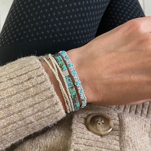 May include: A close-up of a person's wrist wearing a stack of bracelets. The bracelets are made of silver beads and turquoise stones. The person is wearing a brown sweater.