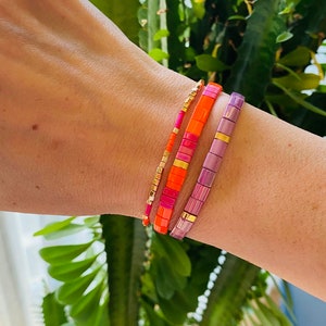Könnte beinhalten: Drei bunte Perlenarmbänder, die auf einem Handgelenk übereinander liegen. Das obere Armband ist helllila mit goldenen Akzenten. Das mittlere Armband ist leuchtend orange mit rosa und goldenen Akzenten. Das untere Armband ist eine dünne goldene Kette.