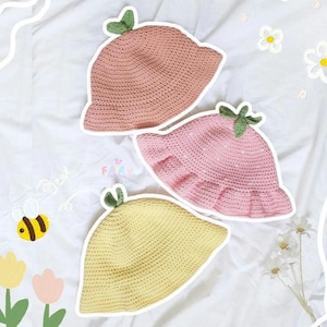 Puede incluir: Tres sombreros de cubo de crochet en rosa y amarillo con detalles de hojas verdes. Los sombreros están sobre un fondo de tela blanca.